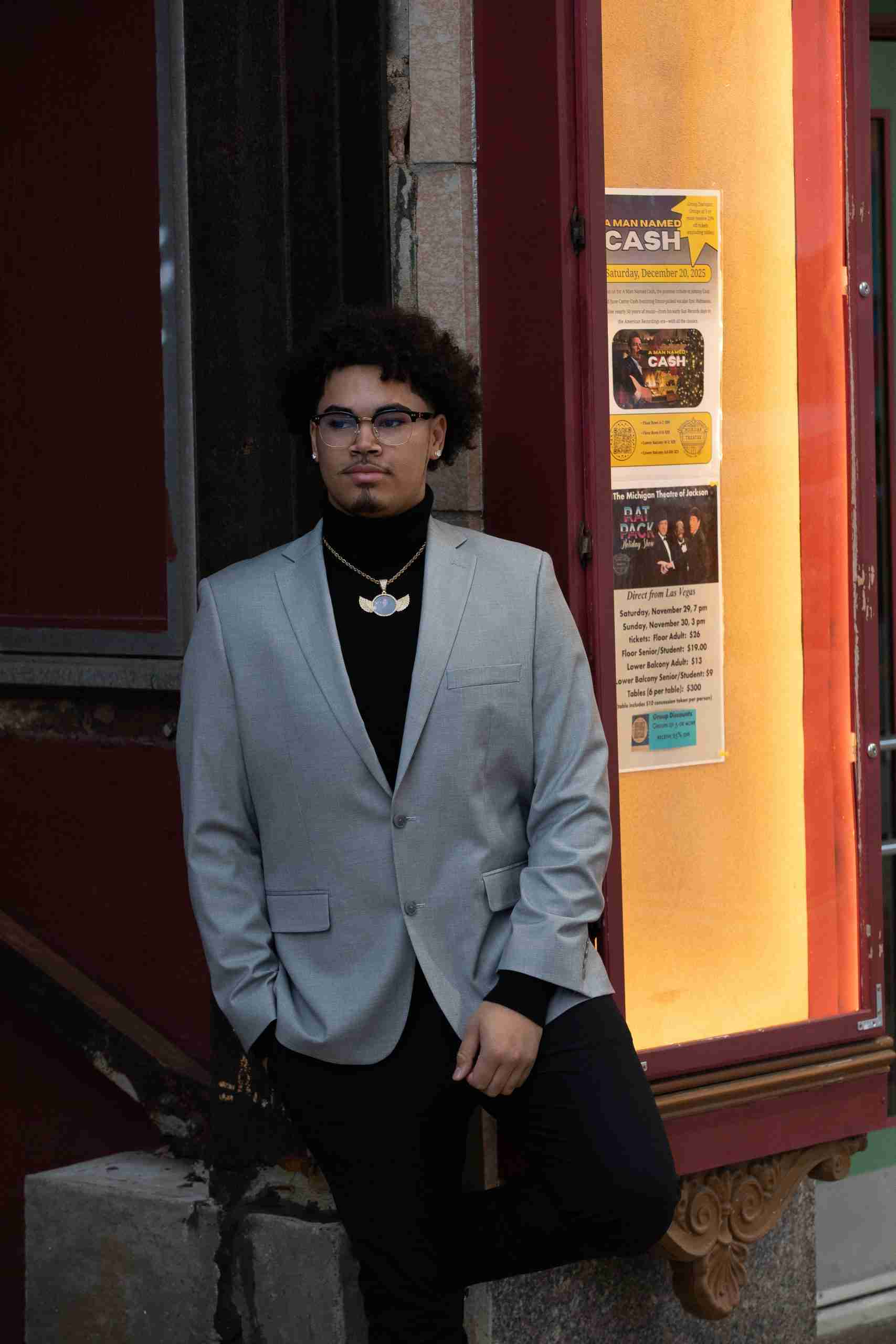 High school senior in glasses and a leather jacket outside an illuminated downtown theater entrance.