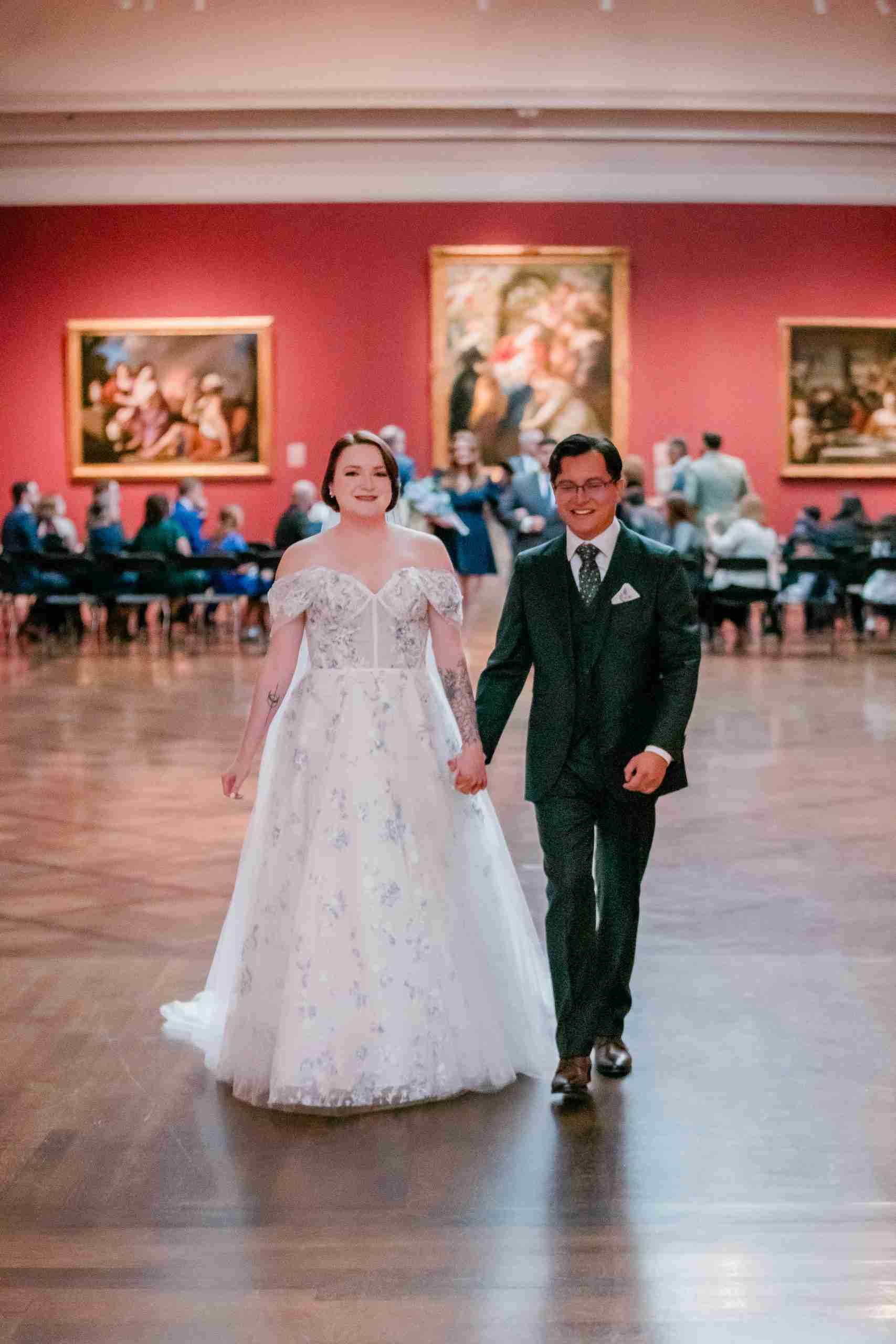 Newlyweds walk hand in hand through an art museum gallery beneath a large painting.